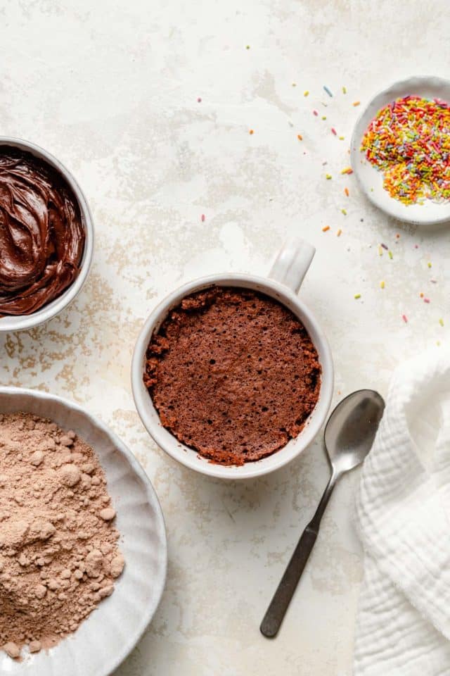 chocolate cake mix mug cake with a bowl of frosting and sprinkles nearby
