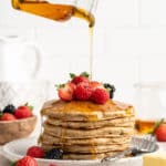 Pouring maple syrup over a stack of whole wheat pancakes with berries.