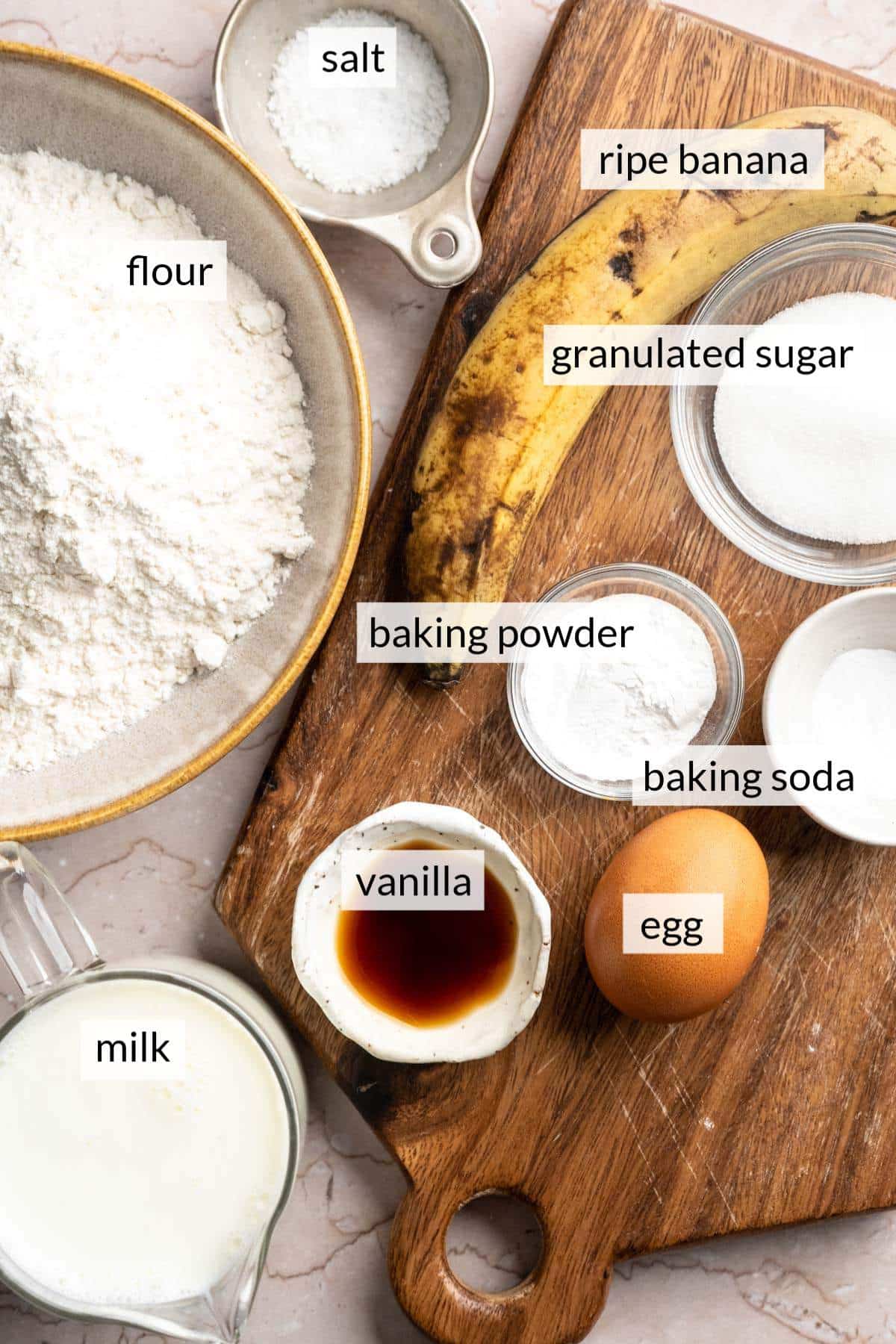 A ripe banana on a cutting board near an egg and bowls with sugar, milk and flour.