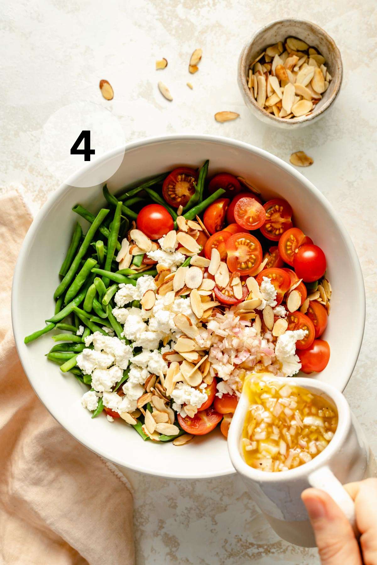 Pouring dressing over green beans with tomatoes, feta and almonds.