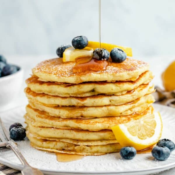 Pouring syrup over stack of pancakes topped with fresh blueberries and served with lemon slices.