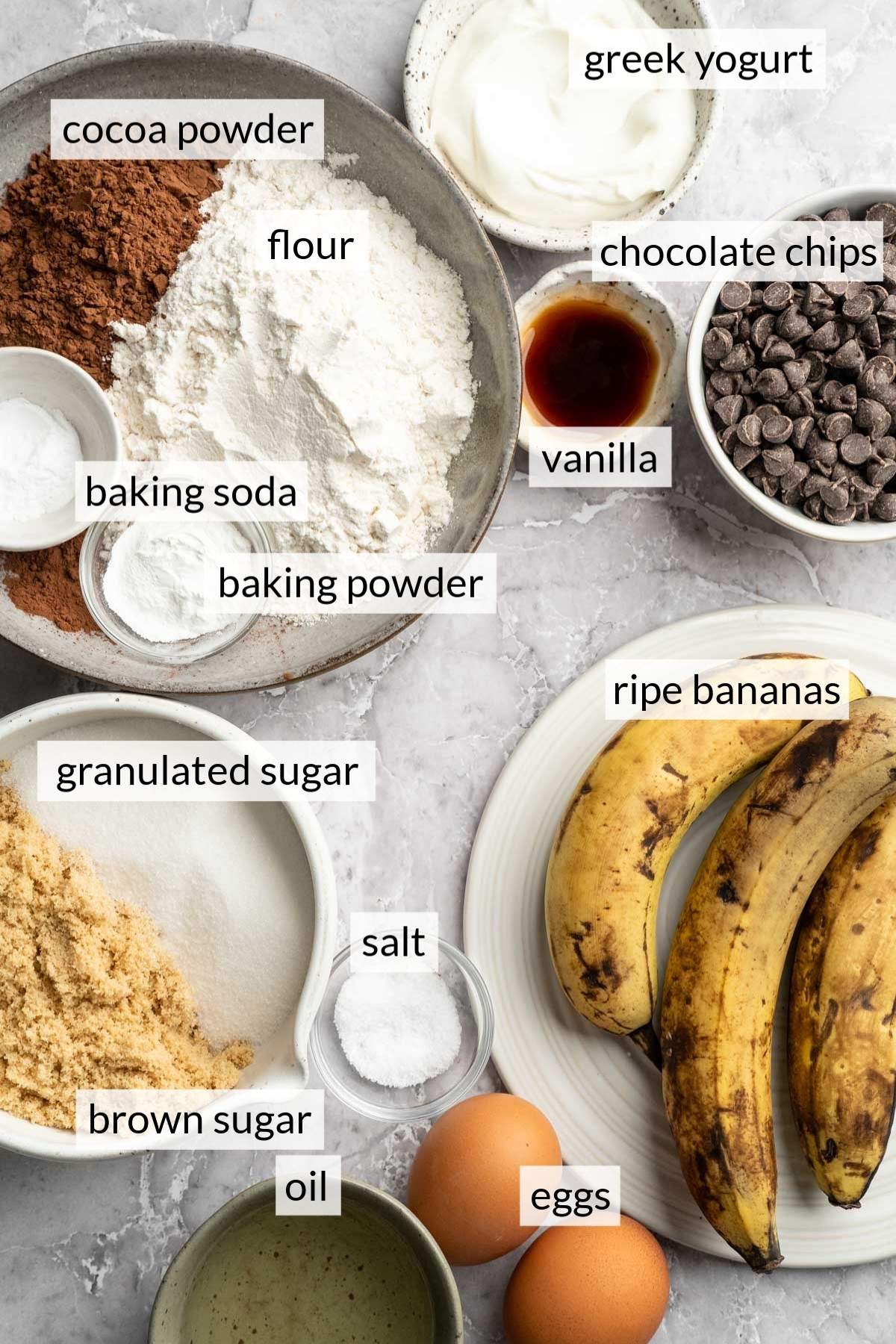 Ripe bananas near eggs and bowls of chocolate chips, Greek yogurt, flour and sugar.