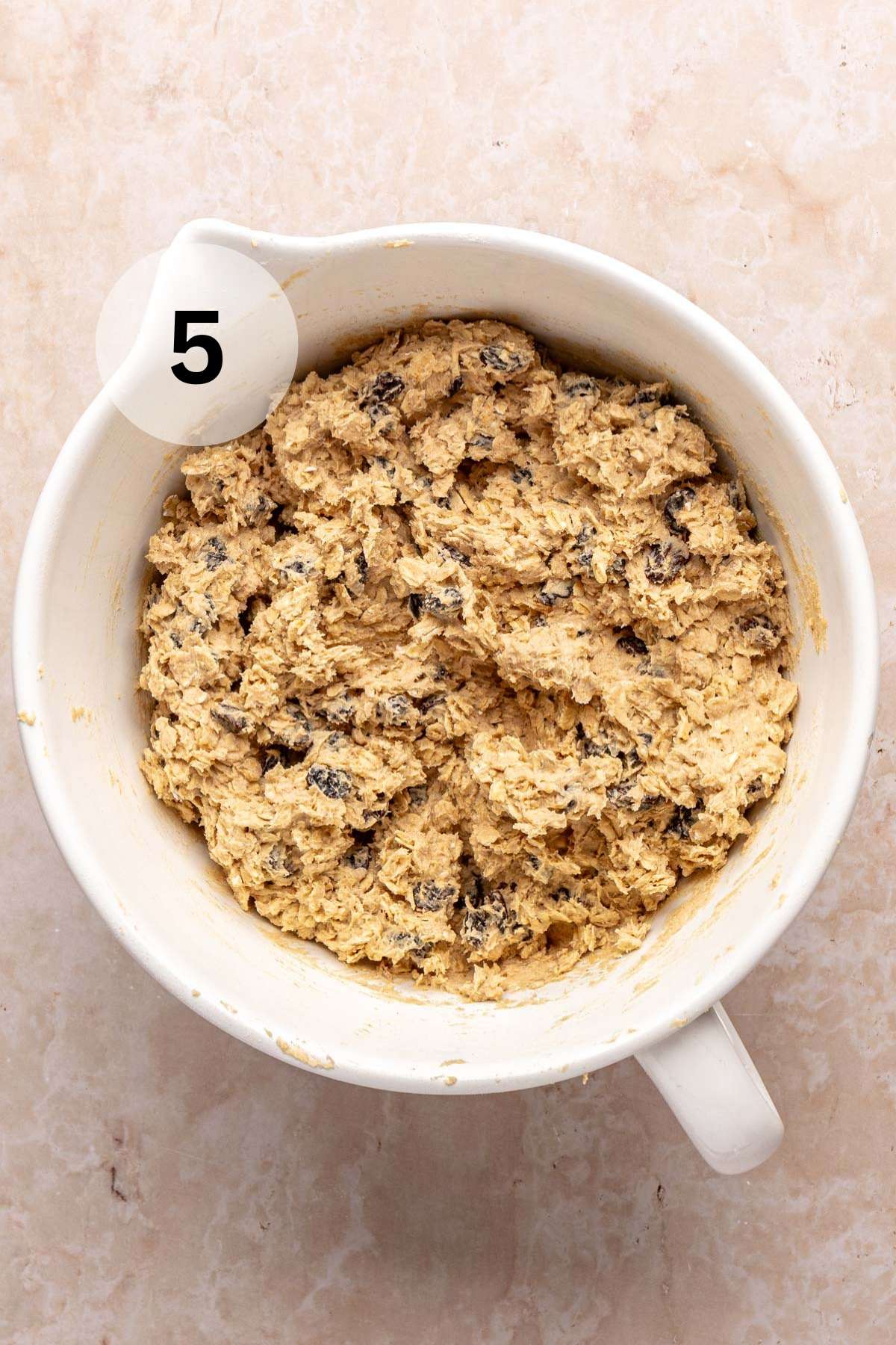Raisins and oats added to cookie dough in a white mixing bowl.