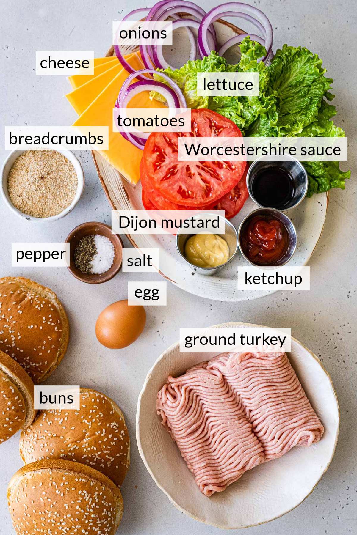 Raw ground turkey in a bowl near hamburger buns, and toppings like cheese, tomatoes and lettuce.