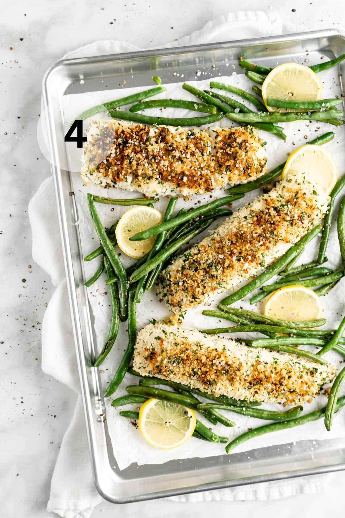 Baked halibut fillets on a baking sheet with green beans and lemon slices.