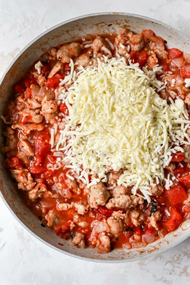 Adding shredded cheese to a tomato sausage mixture in a skillet.