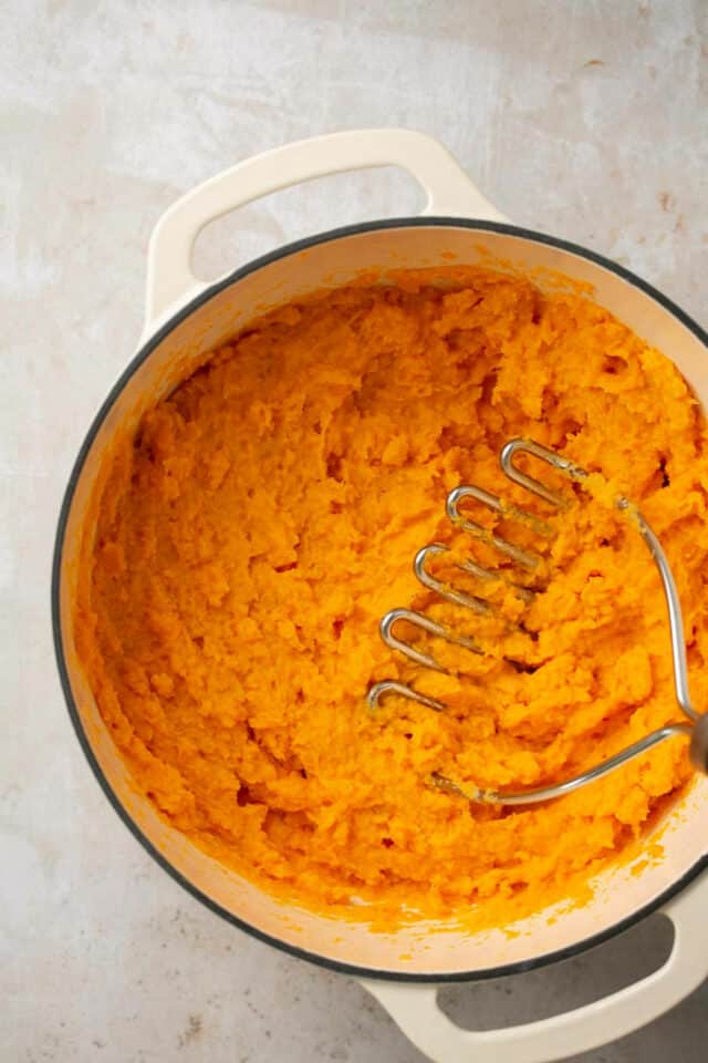 Mashing sweet potatoes with a metal potato masher in a large pot.