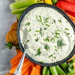Fresh cut veggies and crackers around a bowl of dip with herbs on top.