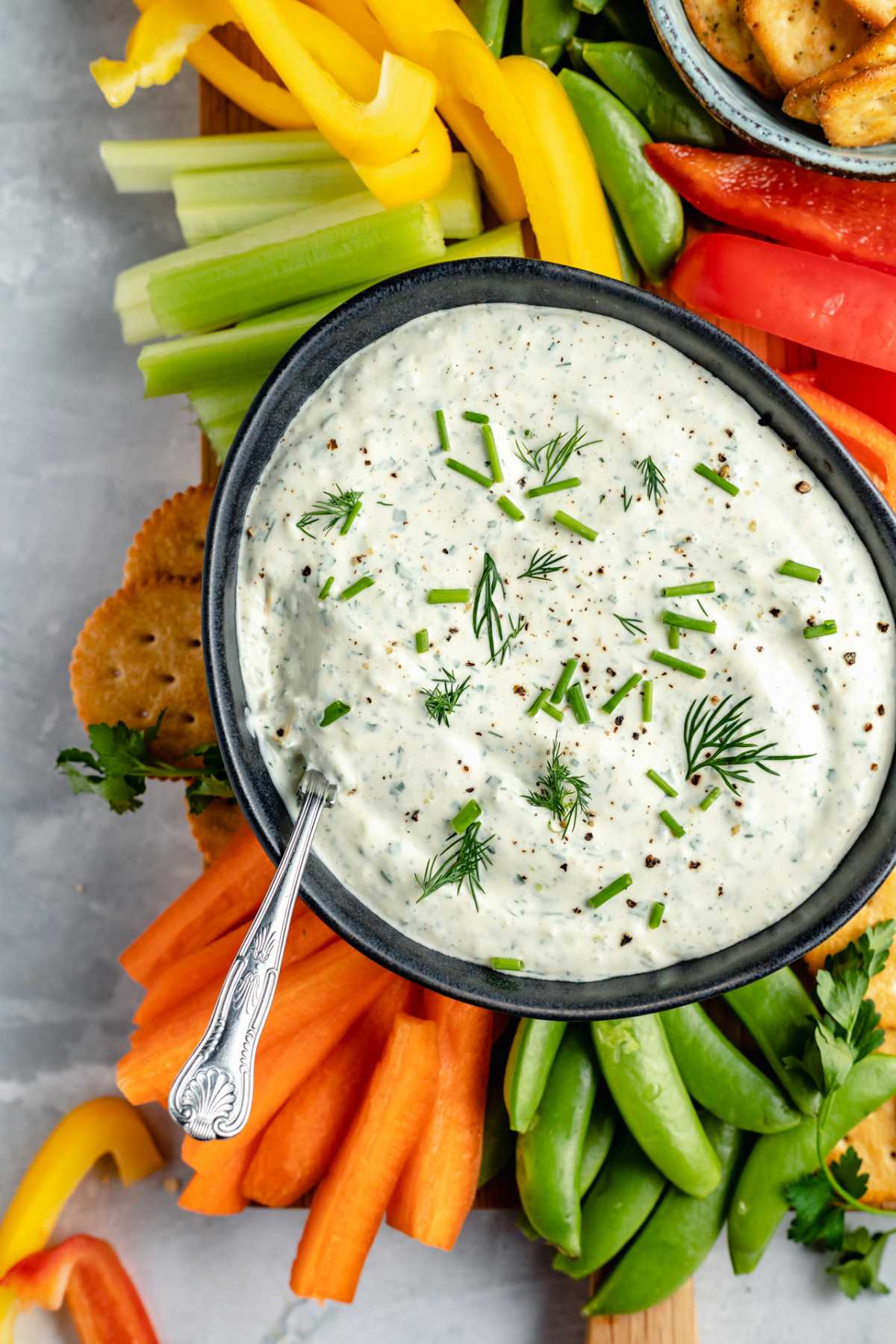 Fresh cut veggies and crackers around a bowl of dip with herbs on top.