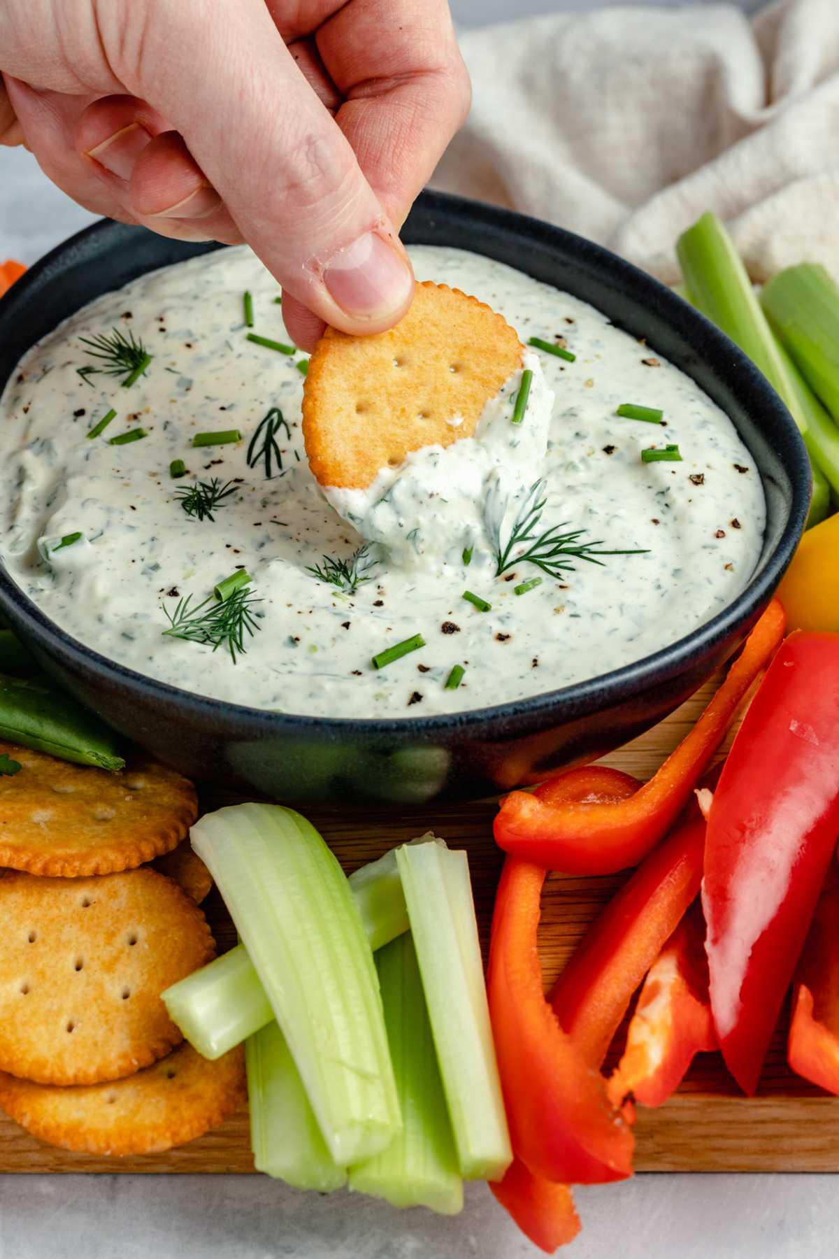 Hand dipping a cracker into a creamy dip with fresh herbs.