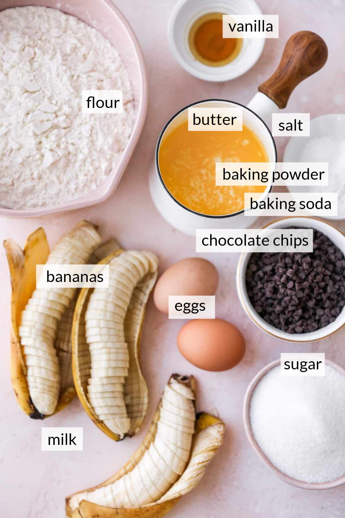 Sliced bananas near bowls of sugar, melted butter, flour, vanilla and chocolate chips.