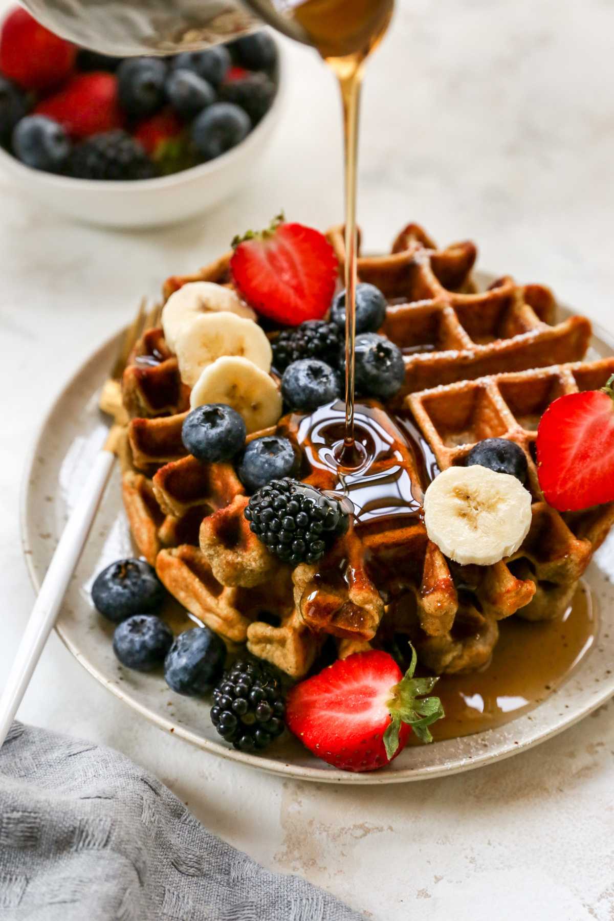 Drizzling maple syrup over a stack of waffles topped with fruit.