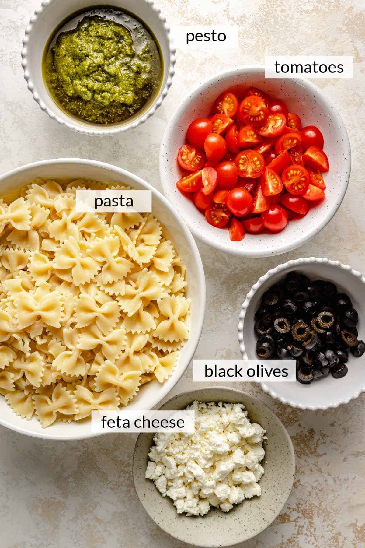 Pasta, feta cheese, black olives, cherry tomatoes and pesto divided into small bowls.