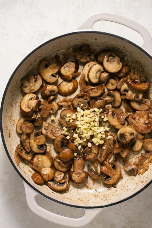 Adding minced garlic to cooked mushrooms in a pan.