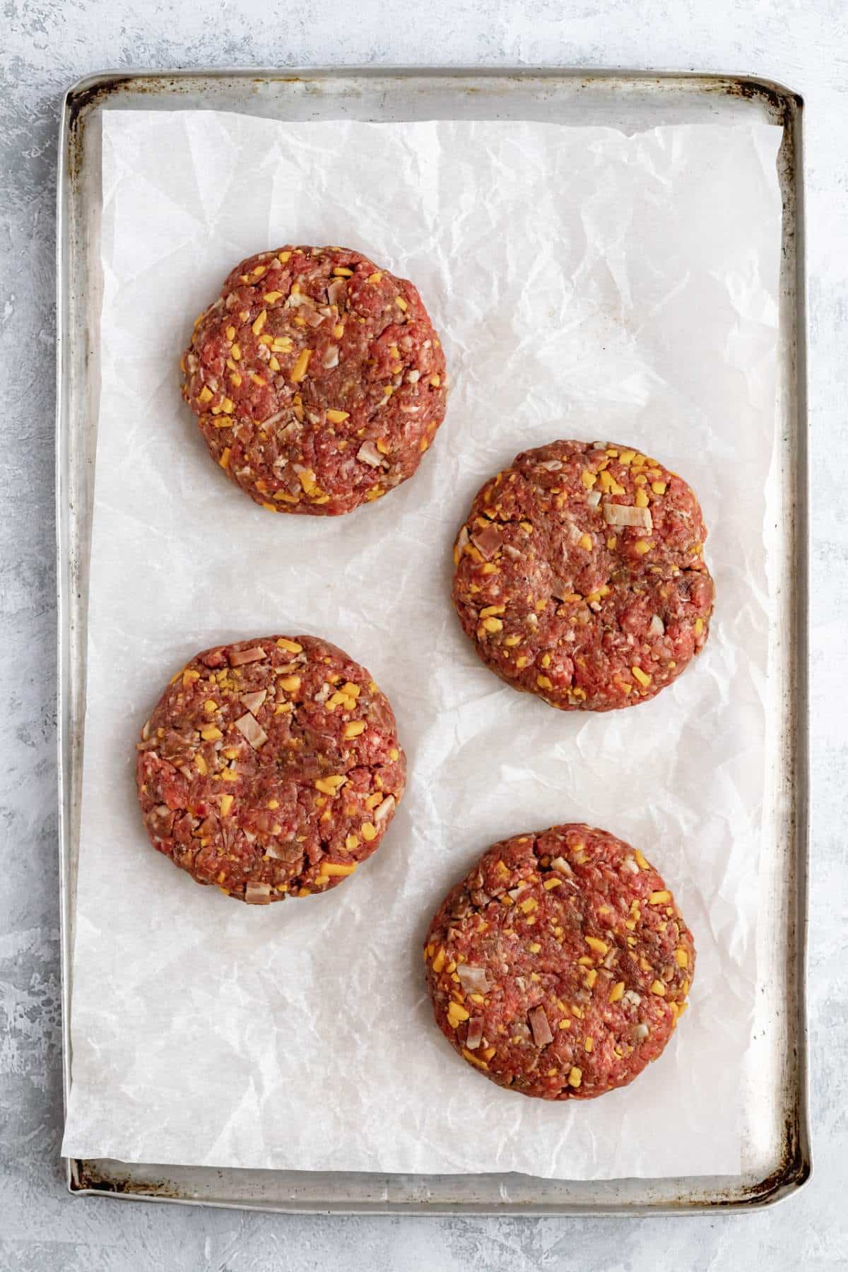 Raw beef patties made with cheese and bacon on parchment paper.