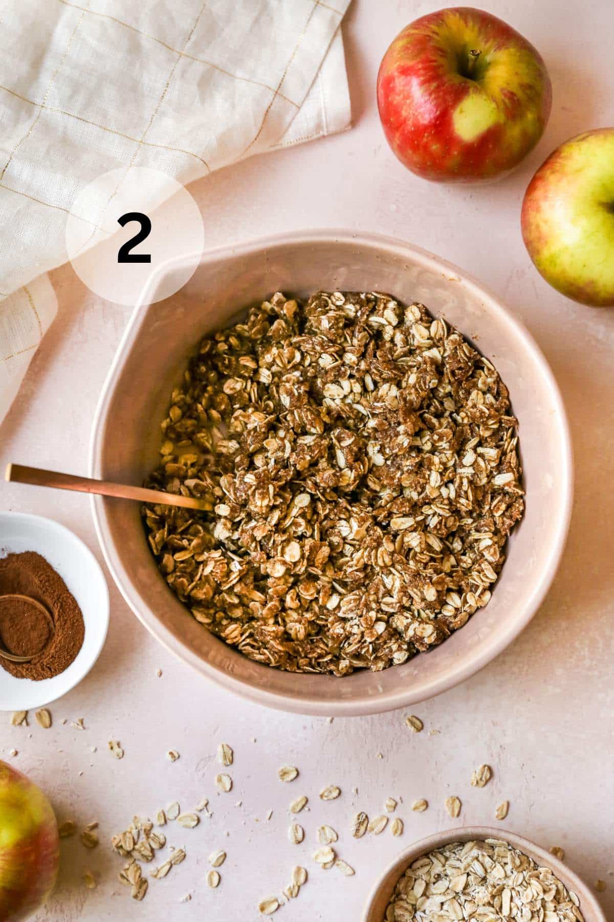 Oat mixture in a large bowl near apples and a small bowl of cinnamon.