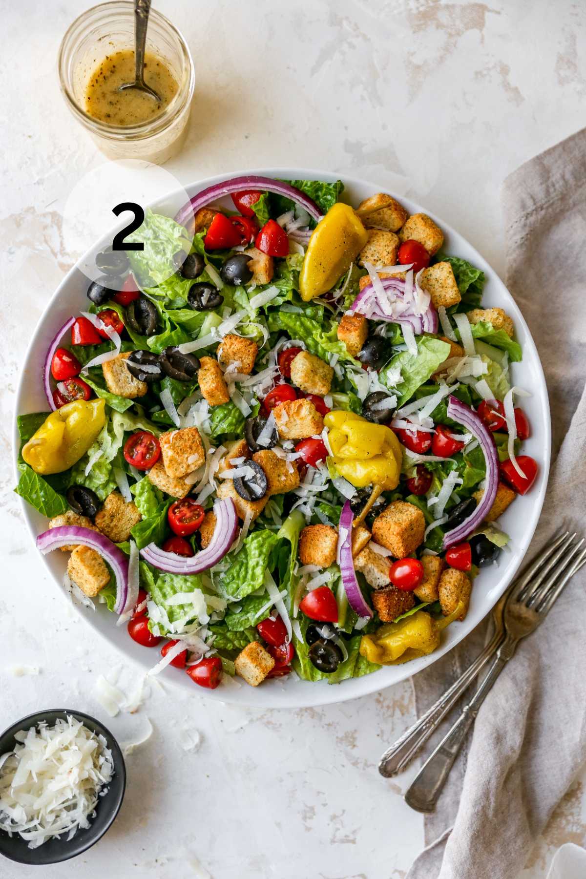 Italian salad in a large white salad bowl near parmesan cheese and dressing.