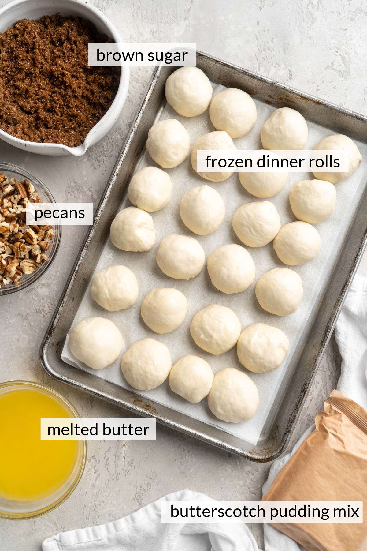 Frozen dinner rolls on a baking sheet pan near pudding mix, melted butter, brown sugar and chopped pecans.
