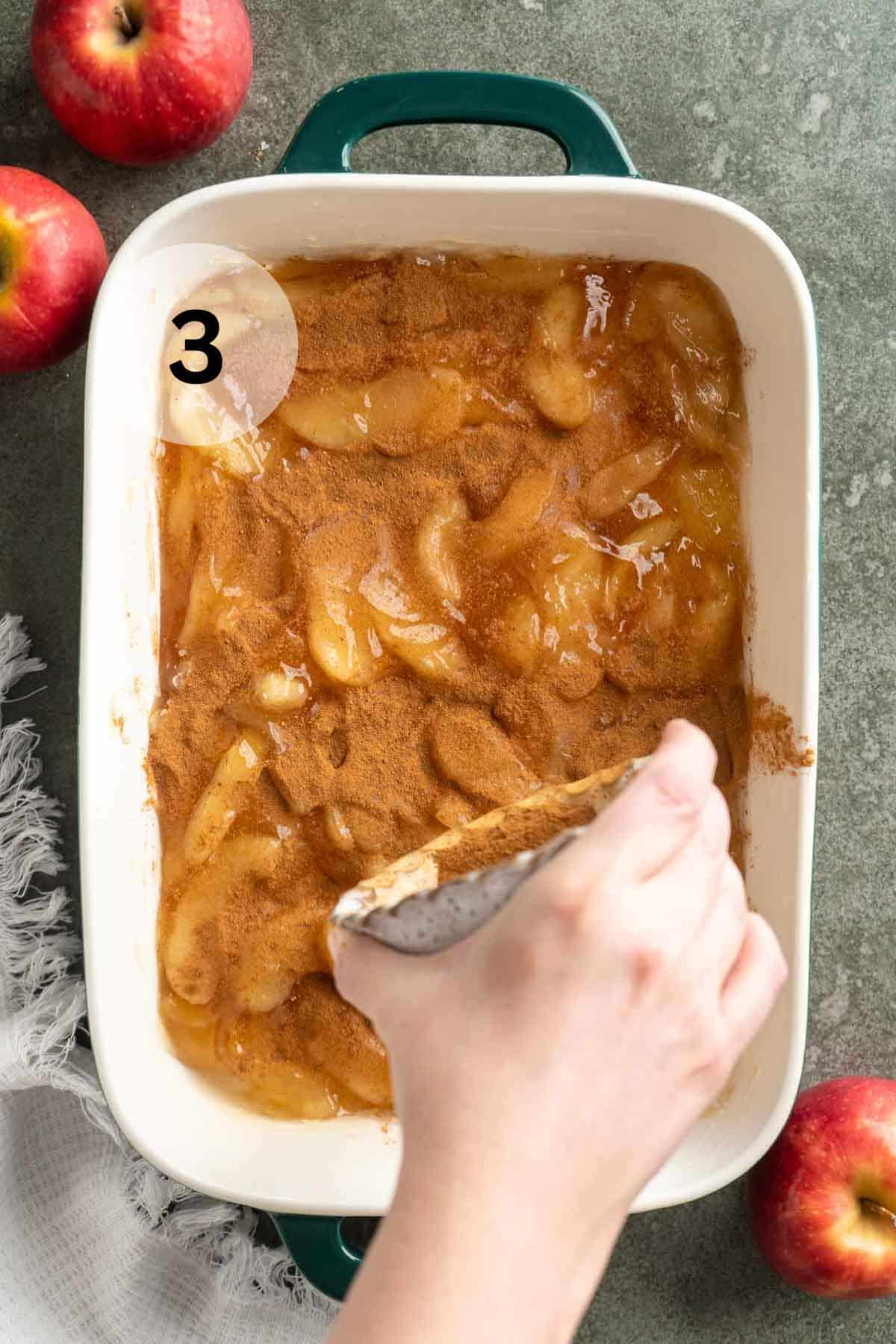 Hand holding a small bowl of spices over sliced apples and sprinkling the spices over the top.