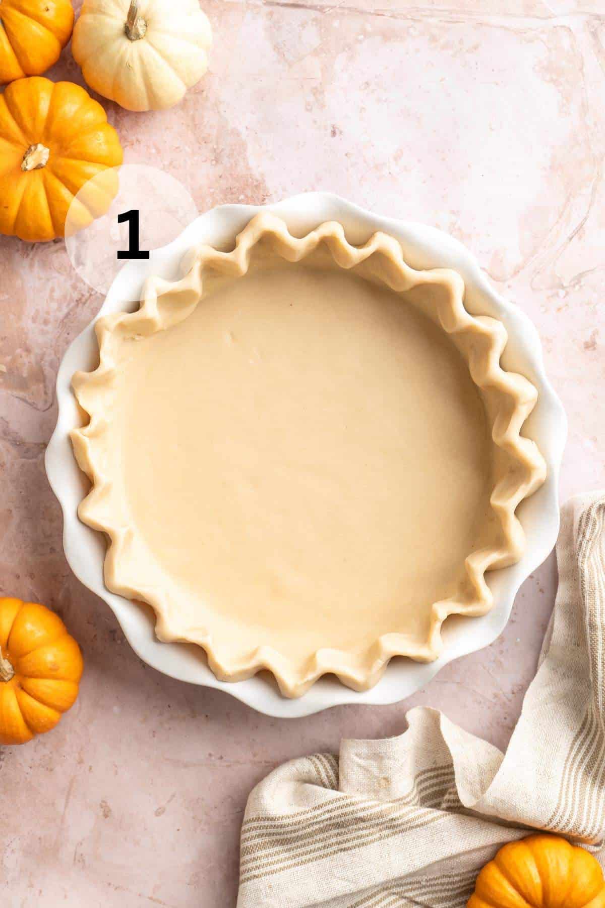 Pie crust in a pie plate near small pumpkins.