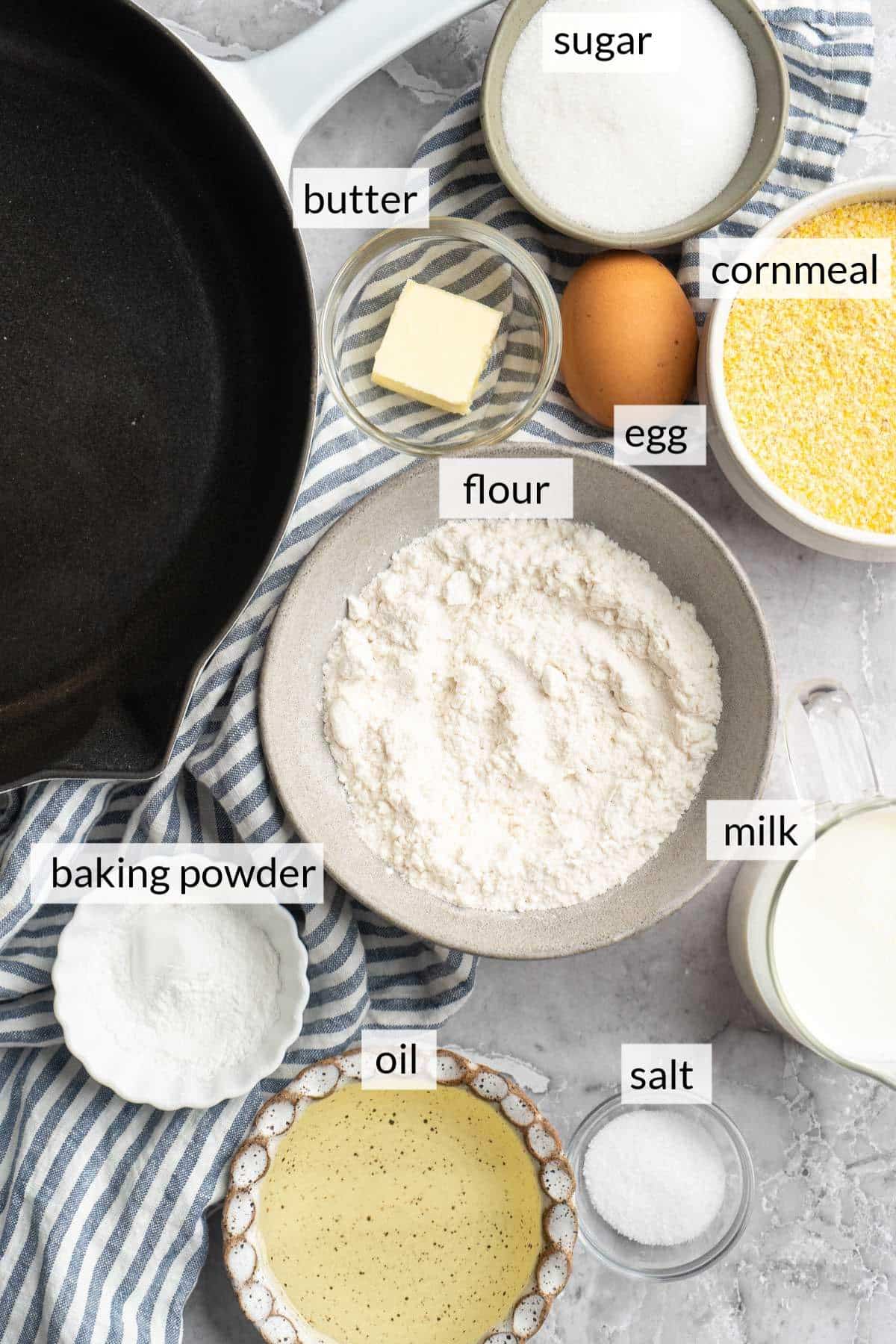 Small bowls with flour, milk, sugar, cornmeal, oil and baking powder near an iron skillet.