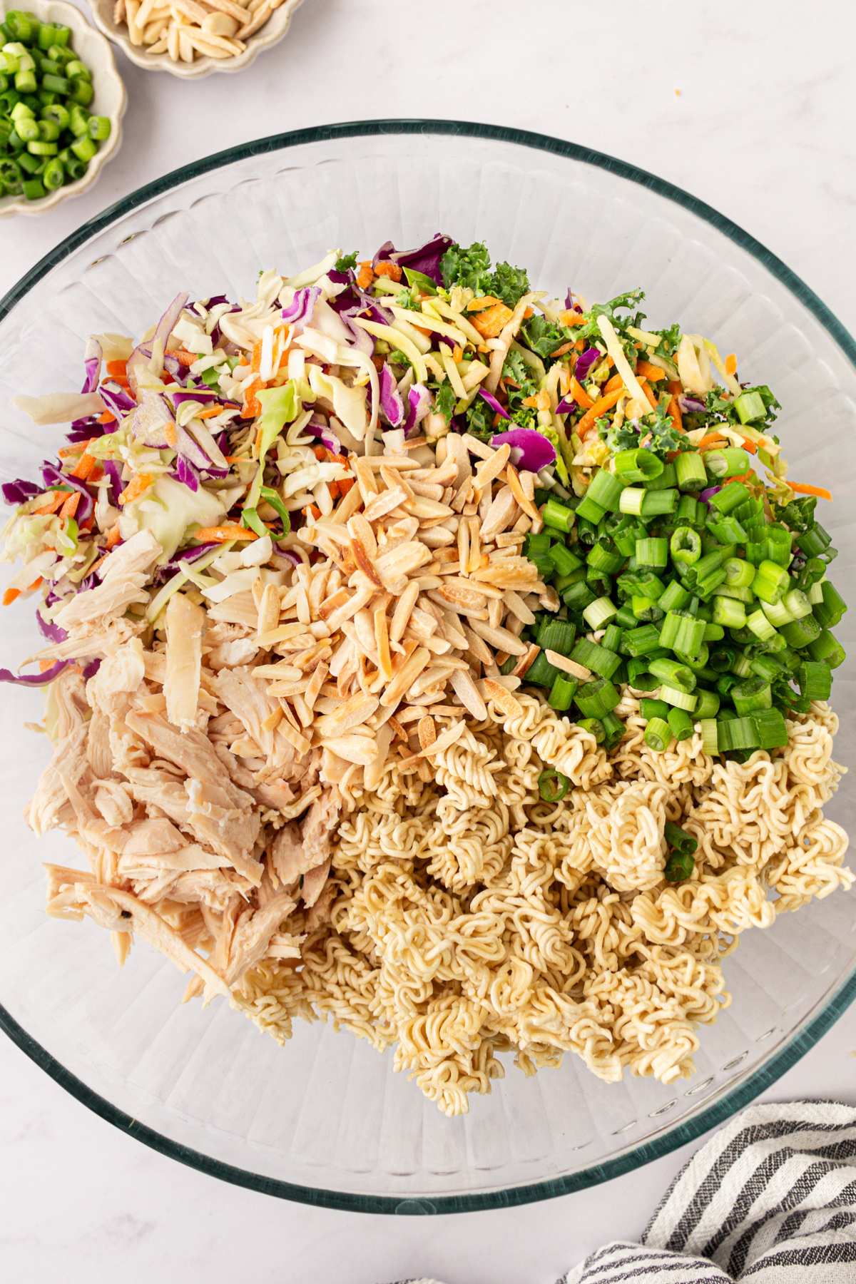 Large bowl with uncooked ramen noodles, green onion, shredded chicken, cabbage and broccoli slaw.