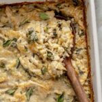 Serving chicken orzo bake from a white dish with a wooden spoon.