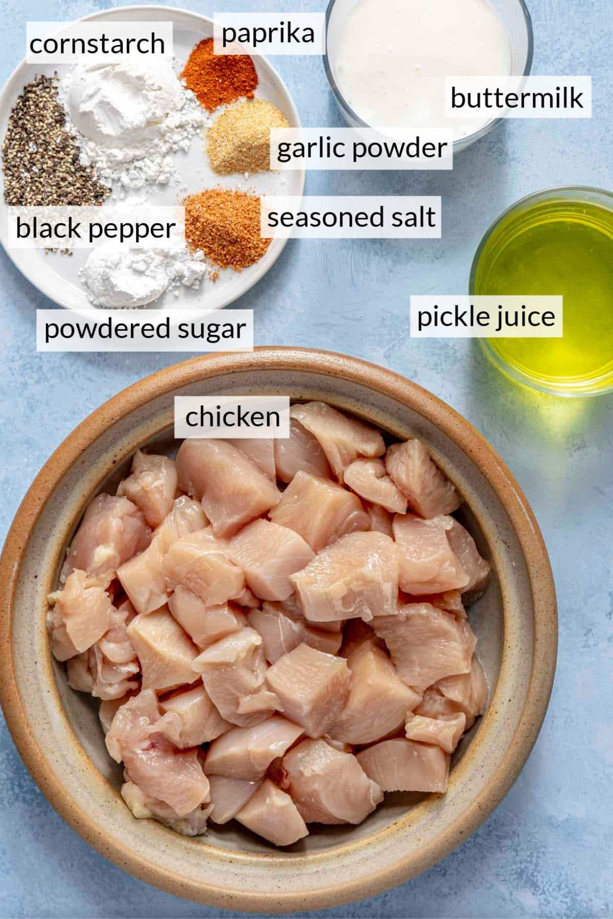 Raw chicken pieces in a bowl near a plate of seasonings, a glass of buttermilk and a glass of pickle juice.