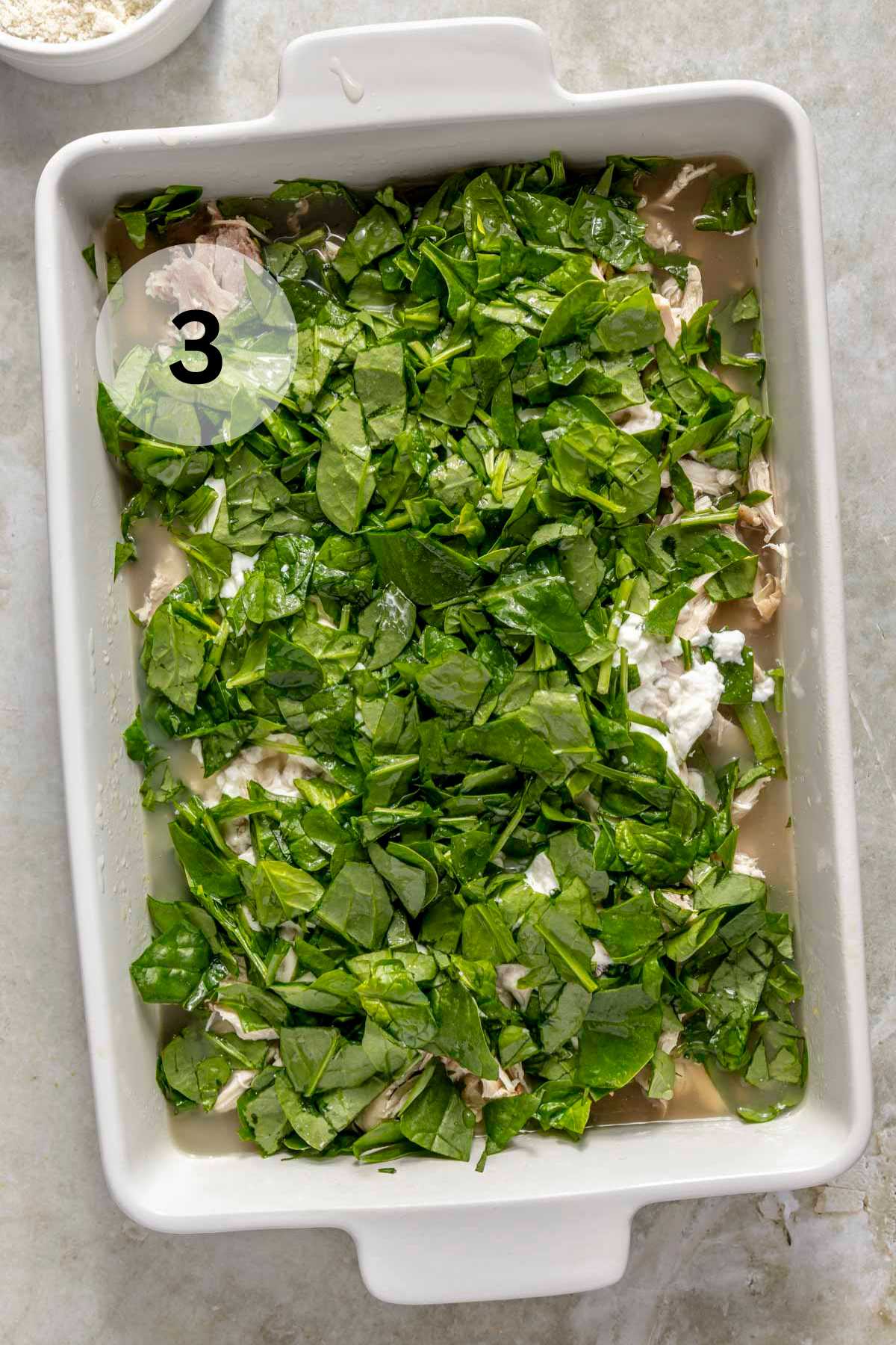 Chopped spinach in a casserole dish with chicken and chicken broth.
