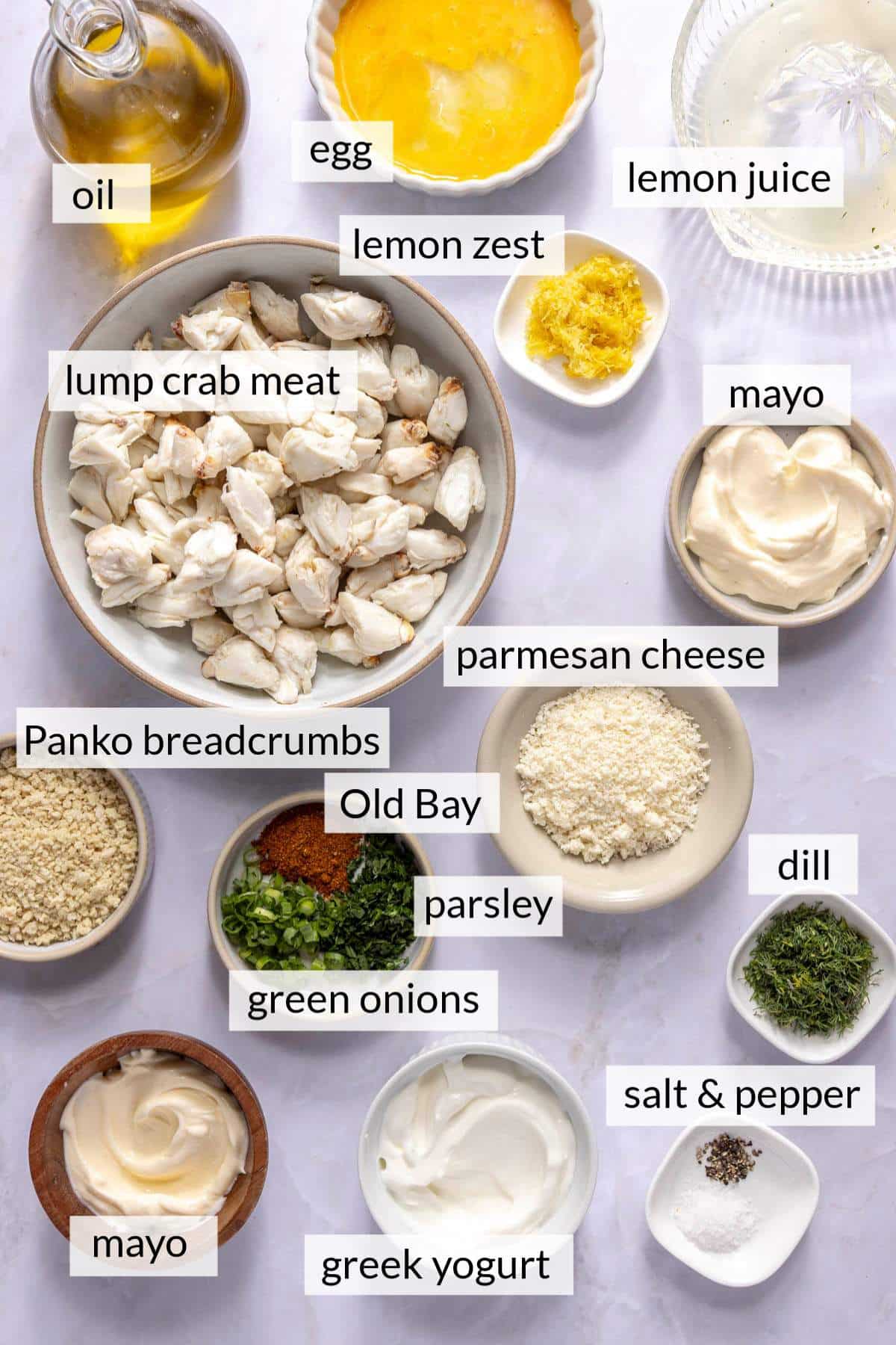 Fresh lump crab meat in a bowl near small bowls of other ingredients like lemon juice, mayo, egg, herbs and breadcrumbs.