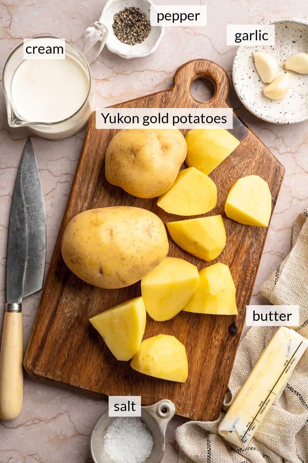 Chopped potatoes on a cutting board near a stick of butter, heavy cream, cloves of garlic and small bowls of salt and pepper.