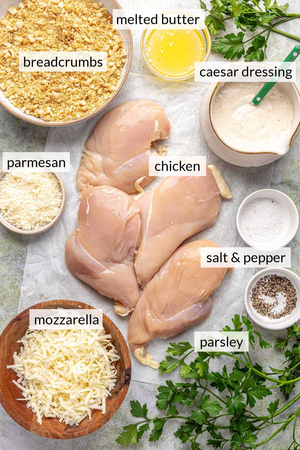 Raw chicken breasts near small bowls of parmesan cheese, breadcrumbs, caesar dressing and melted butter.