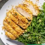 Breaded caesar dressing chicken on a white plate with a simple arugula salad.