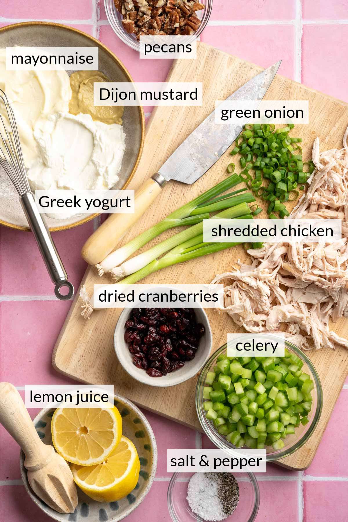 Chopped green onion and shredded chicken on a cutting board near small bowls of lemons, cranberries, celery and mayo.