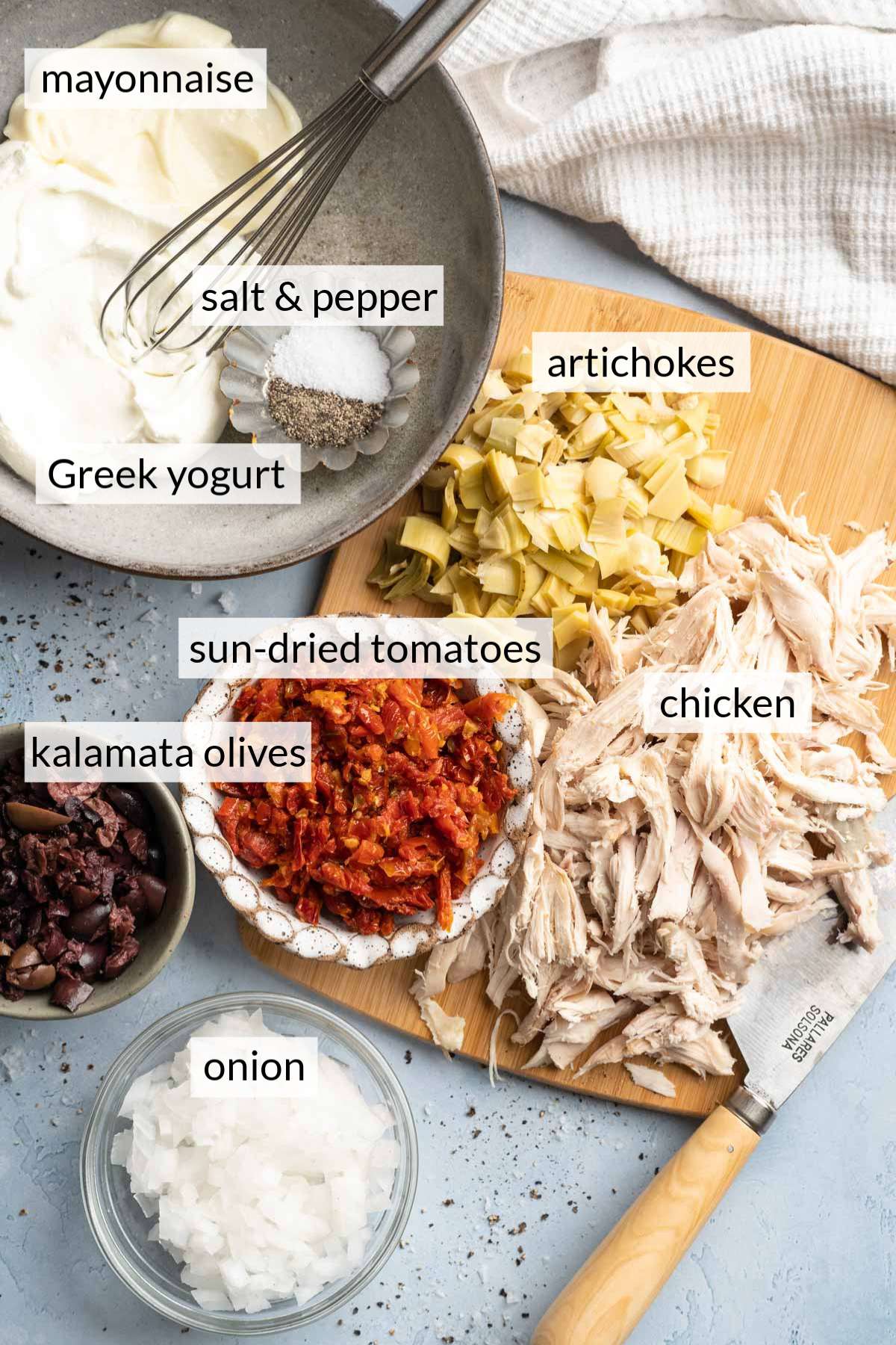 Chopped chicken and artichokes on a cutting board near bowls with onions, olives, mayo and Greek yogurt.