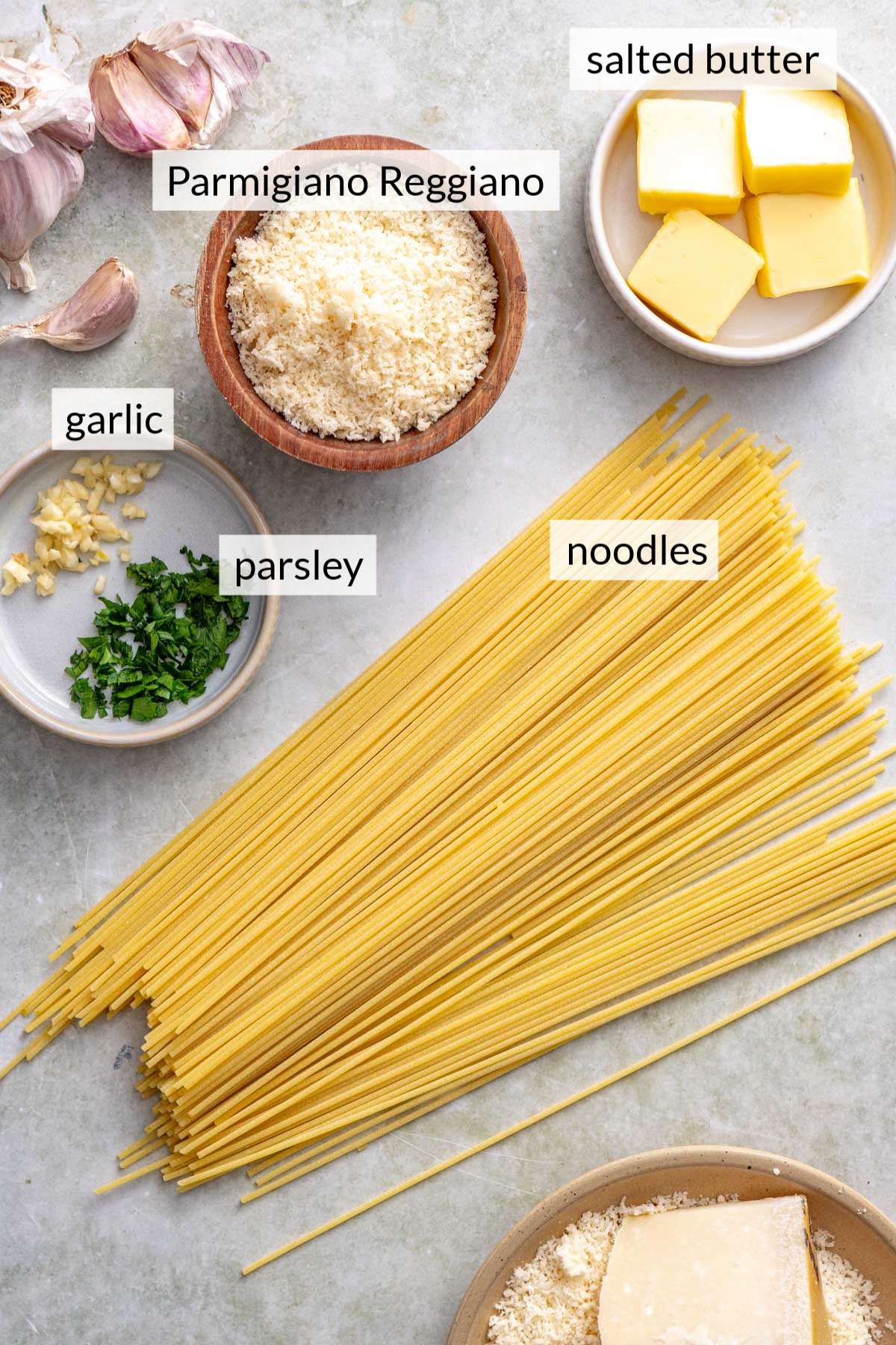 Uncooked spaghetti noodles near bowls of butter, garlic and parmesan cheese.