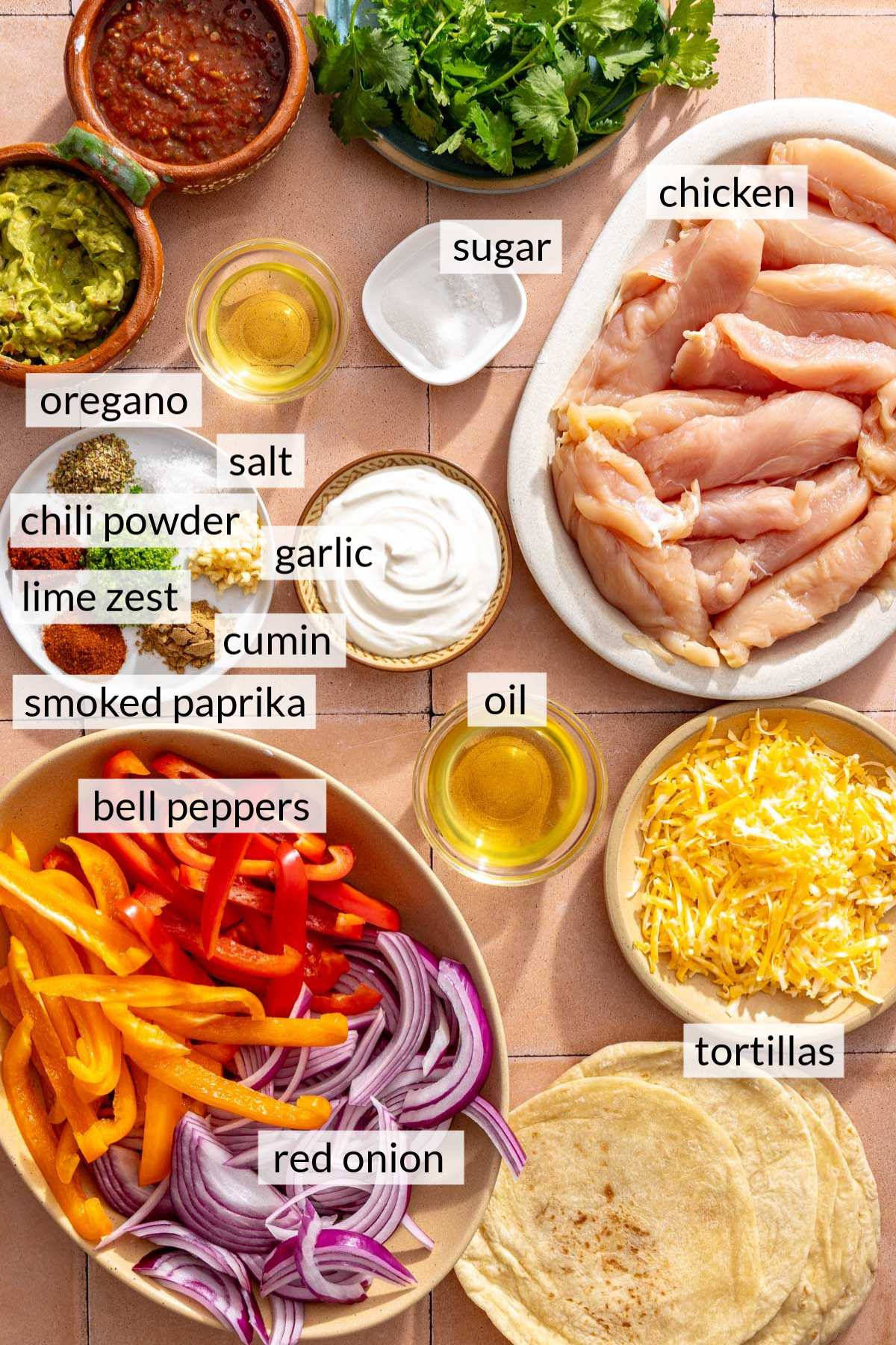 Raw chicken tenderloins near freshly sliced bell pepper and red onion, spices, shredded cheese and tortillas.