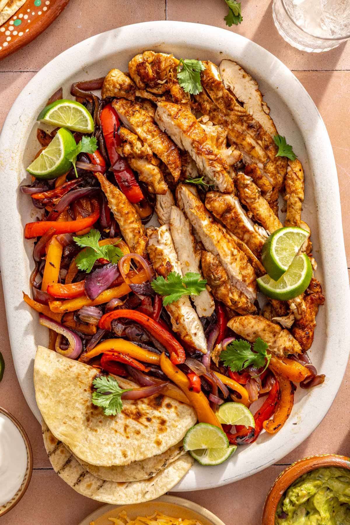Sliced grilled chicken on a platter with charred veggies and tortillas.