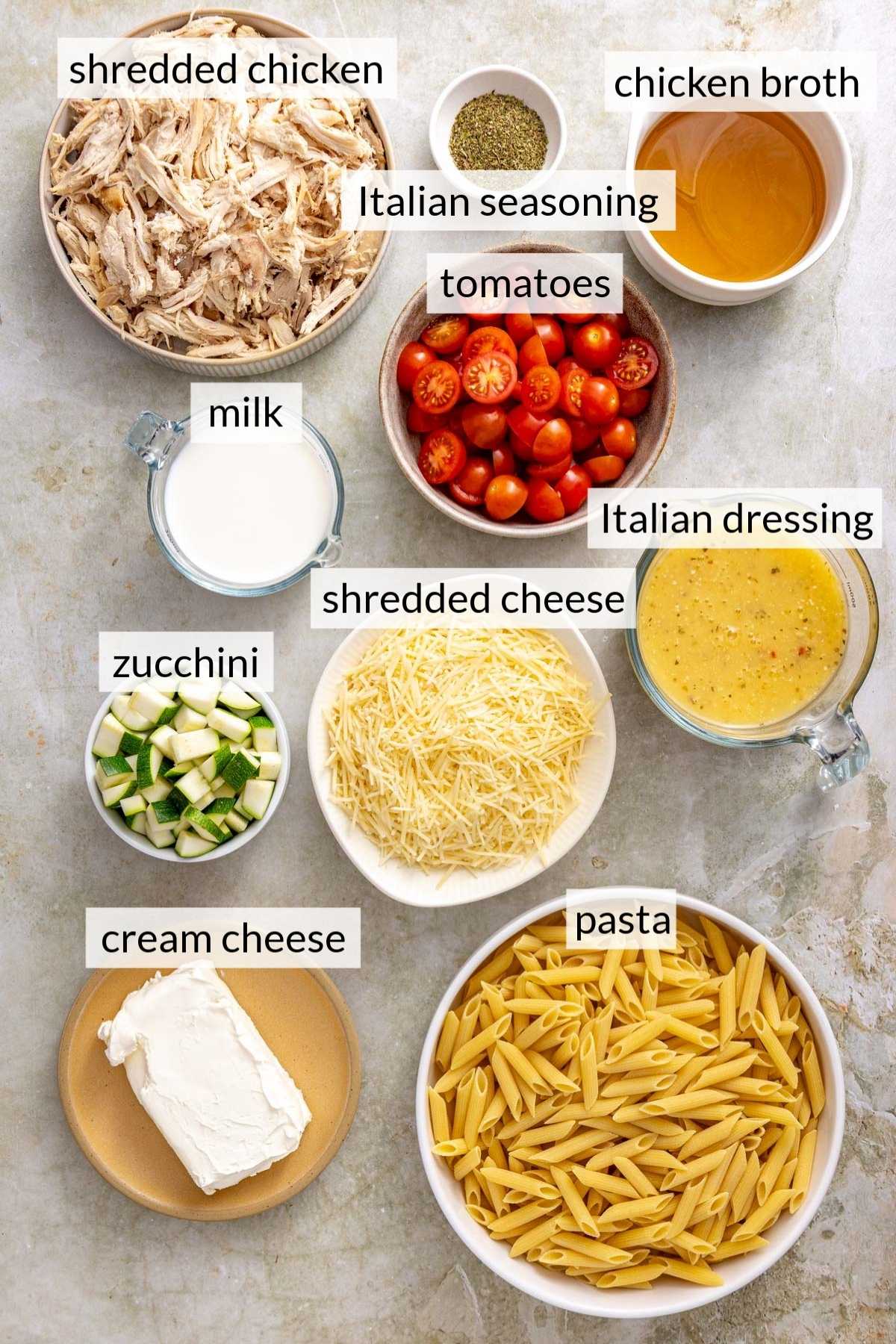 A block of cream cheese near bowls of uncooked pasta, shredded cheese, tomatoes, zucchini, milk and shredded chicken.