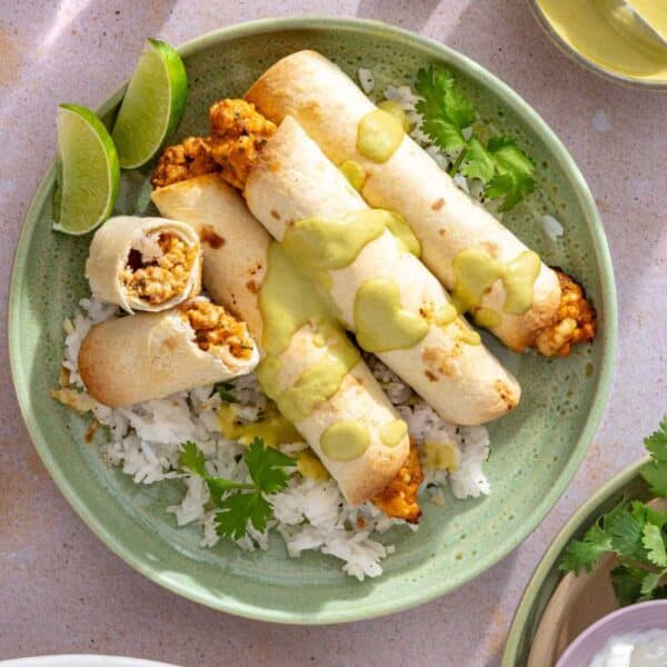 Chicken taquitos drizzled with a creamy green sauce on a plate with rice, cilantro and lime wedges.