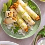 Chicken taquitos drizzled with a creamy green sauce on a plate with rice, cilantro and lime wedges.