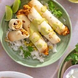 Chicken taquitos drizzled with a creamy green sauce on a plate with rice, cilantro and lime wedges.