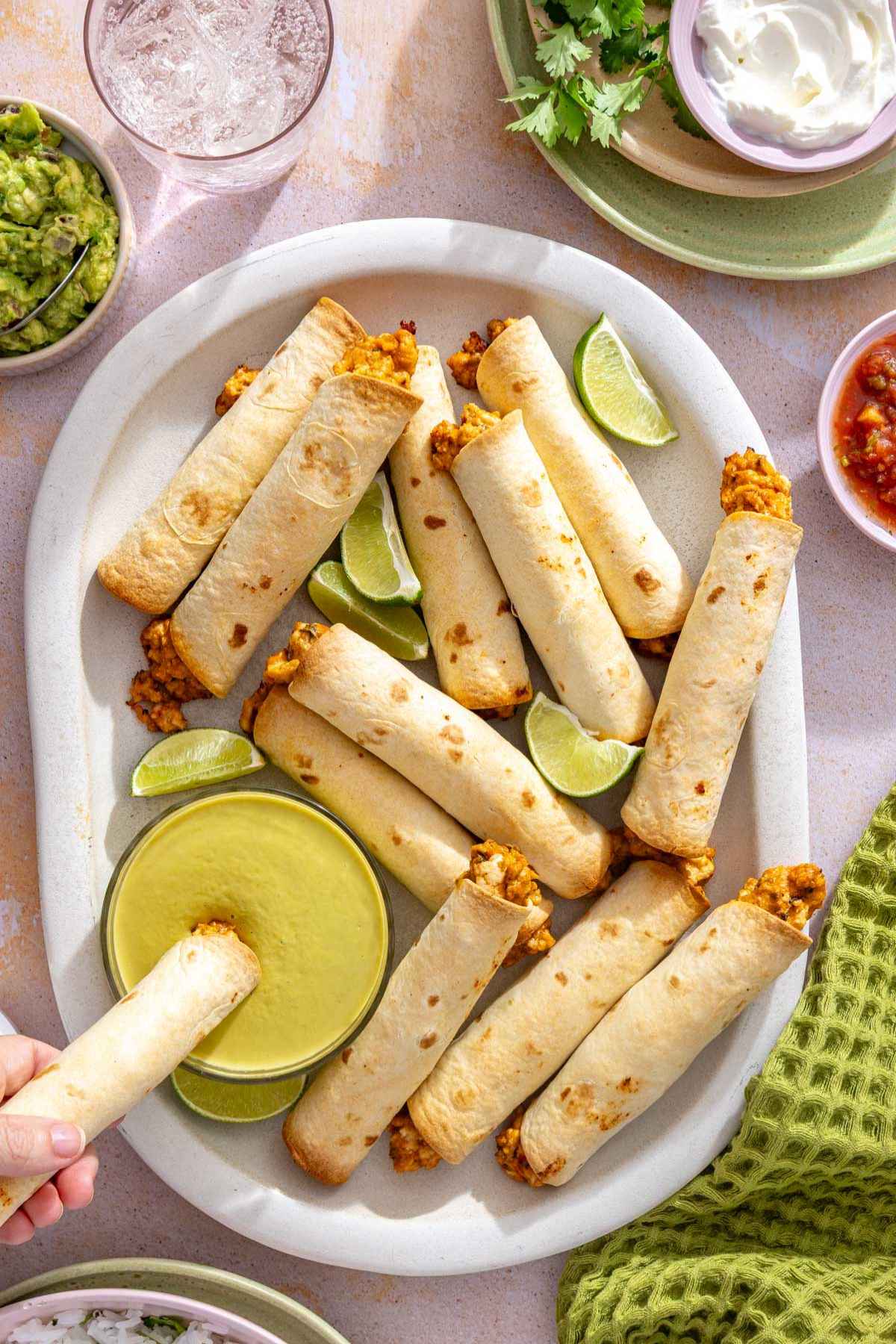 Chicken taquitos on a white serving plate with one being dipped into a small bowl of green sauce.
